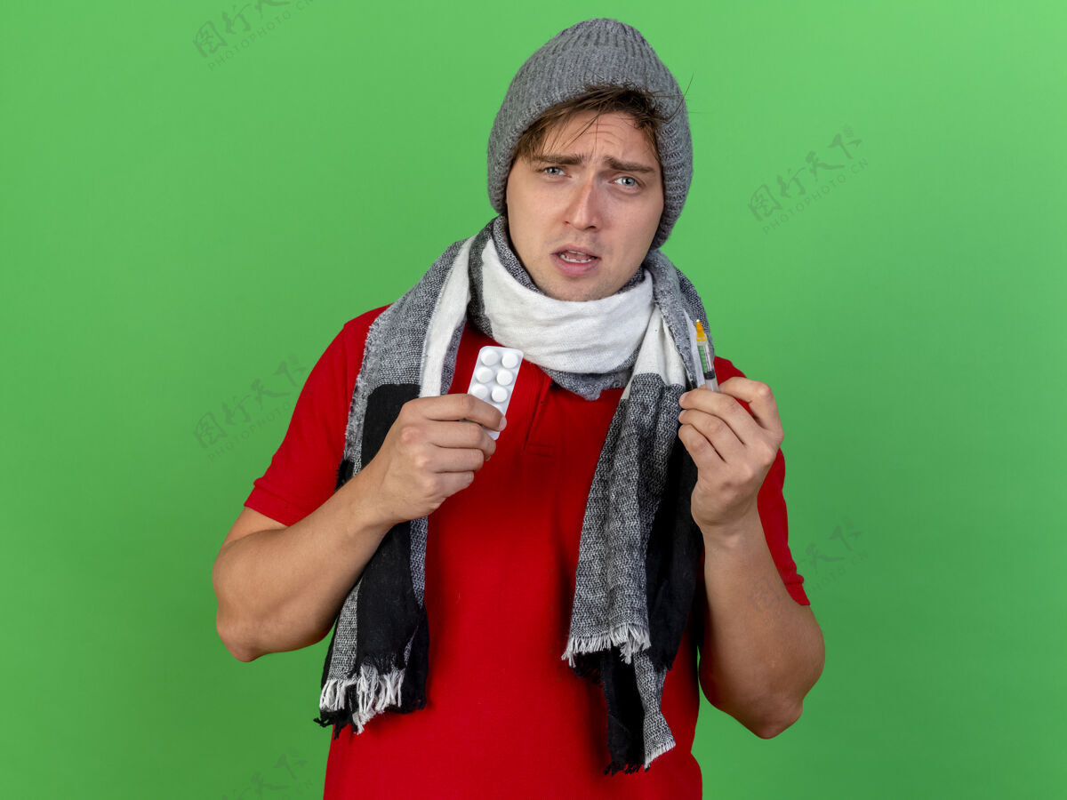 人虚弱的年轻英俊的金发病男子戴着冬天的帽子和围巾拿着一包药片和注射器看着前面隔离的绿色墙壁上的复制空间拿着站着注射器