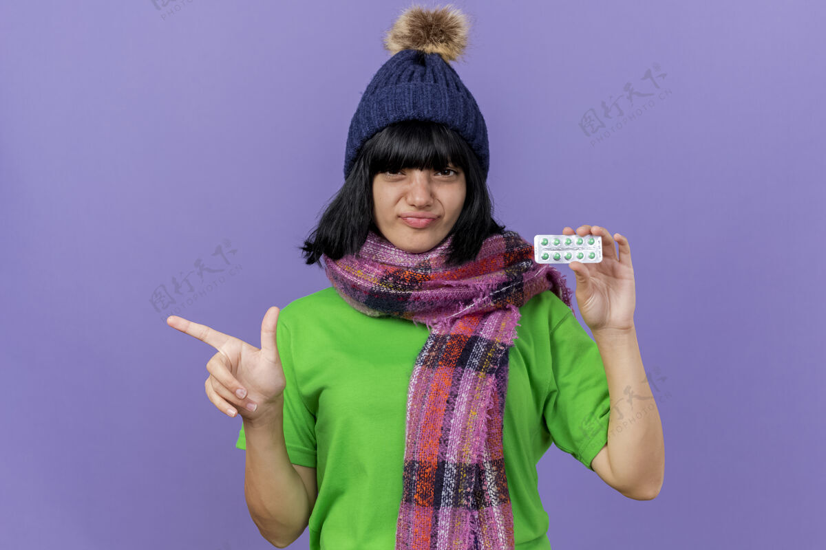 衣服自信的年轻生病的女人戴着冬天的帽子和围巾 看着前面拿着一包胶囊 指着隔离在紫色墙上的一面 留着复印空间信心围巾站