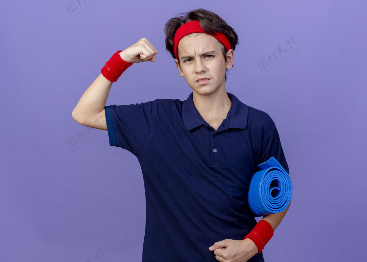 强壮自信的年轻帅气的运动男孩戴着头带和护腕 戴着牙套 拿着瑜伽垫 看着前面做着强烈的手势 孤立在紫色的墙上人站着年轻