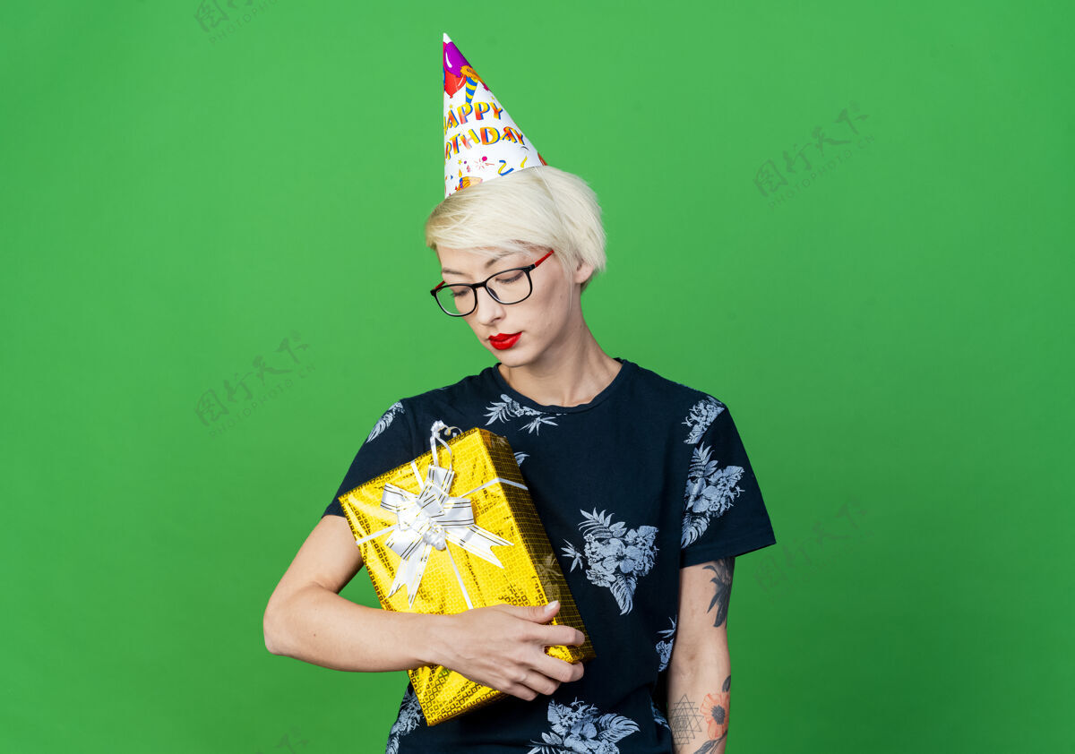 生日年轻的金发派对女郎戴着眼镜 戴着生日帽 拿着礼物盒 看着隔离在绿色墙上的复制空间抱着站着脸