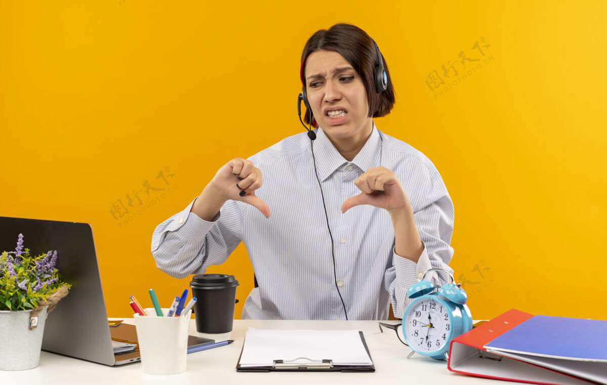 笔记本电脑年轻的电话中心女孩戴着耳机坐在办公桌旁 手里拿着工作工具 看着笔记本电脑 拇指朝下 孤立地站在橙色背景上显示工作不愉快