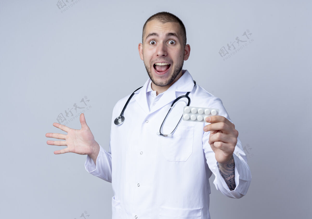 周围令人印象深刻的年轻男医生穿着医用长袍 脖子上戴着听诊器 手里拿着一包药片 一只空手孤零零地站在白纸上空长袍药片