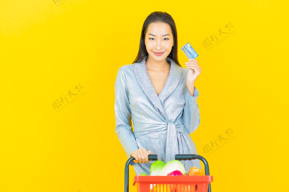 肖像一幅美丽的亚洲年轻女子微笑的画像 黄色的墙上挂着超市的购物篮购物车享受信用卡