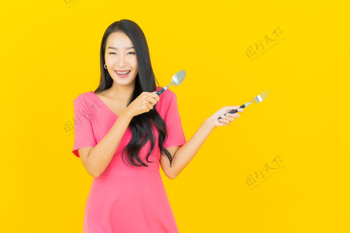 欢呼画像美丽的亚洲年轻女子微笑着用勺子和叉子在黄色的墙上叉子美丽餐具