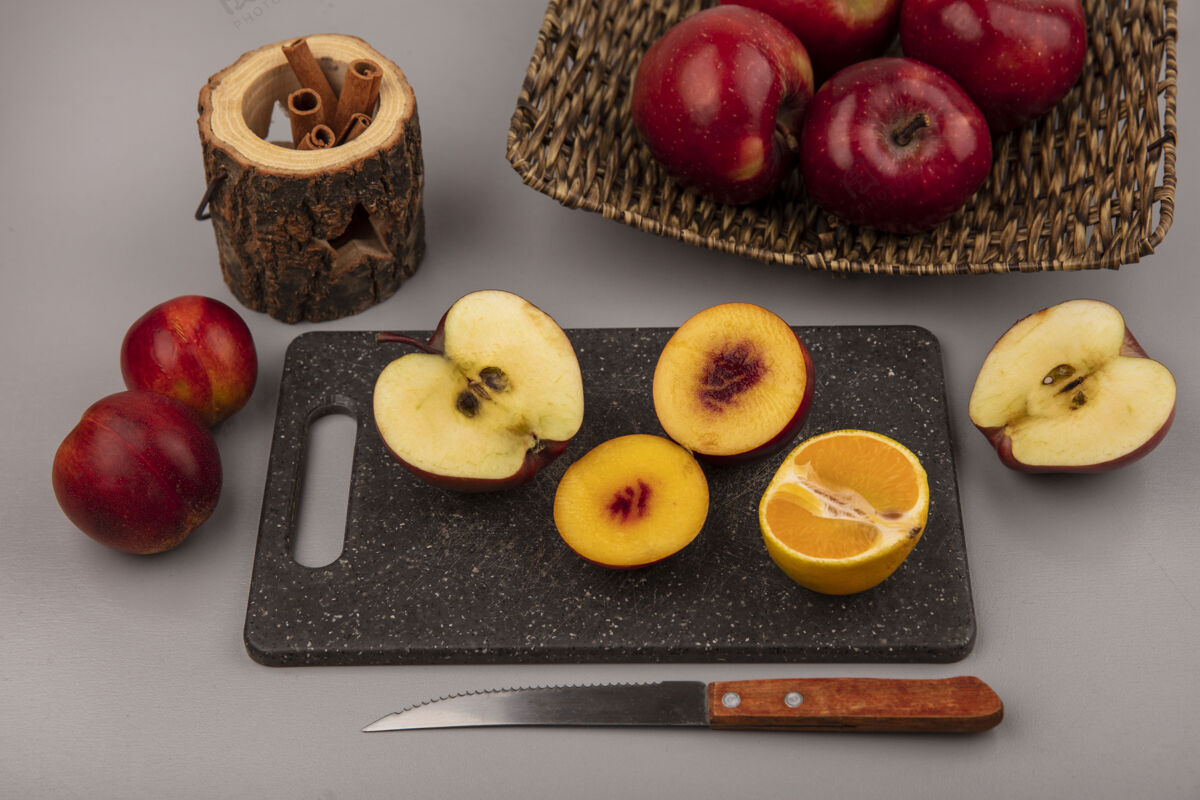 苹果新鲜的半个桃子放在一个黑色的厨房板上 上面有橘子和苹果 刀子上有红苹果 柳条托盘上有一个灰色的背景橘子水果灰色