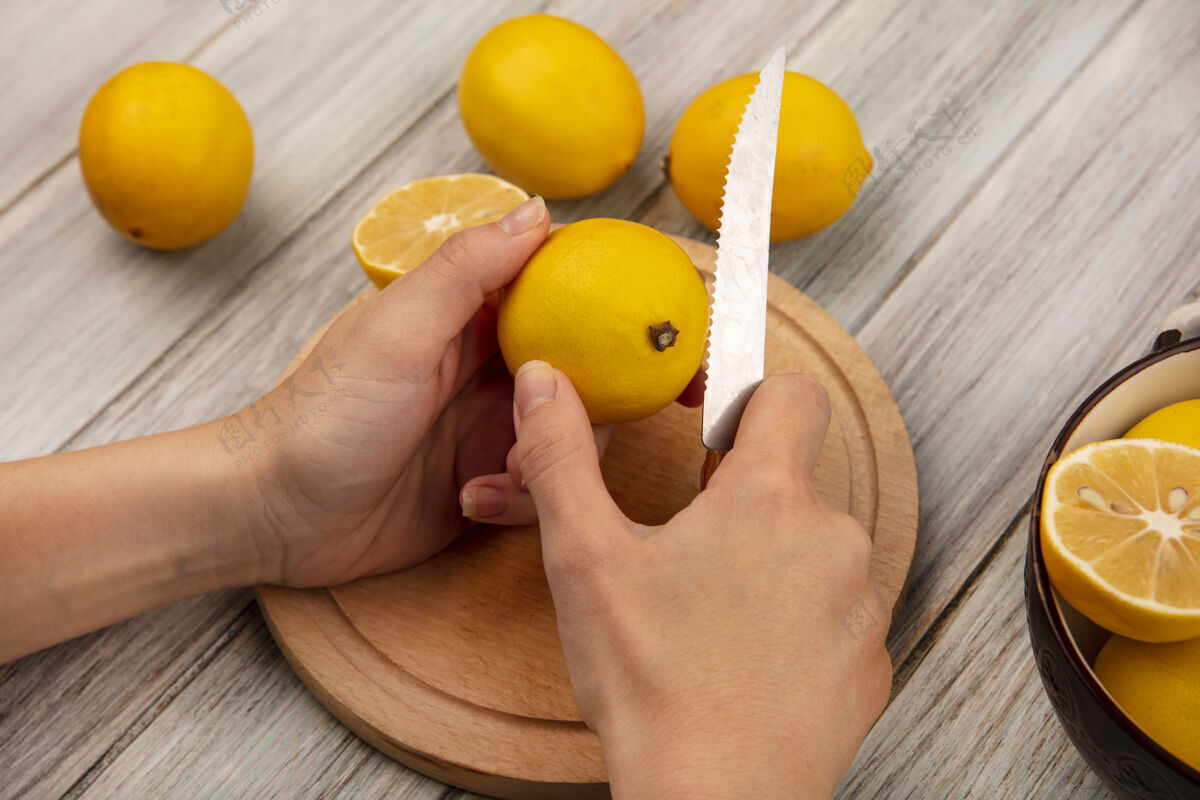 切片女性手在木制厨房板上切割新鲜柠檬的俯视图 刀子上放着柠檬 碗里的柠檬被隔离在灰色的木制背景上健康柑橘食物