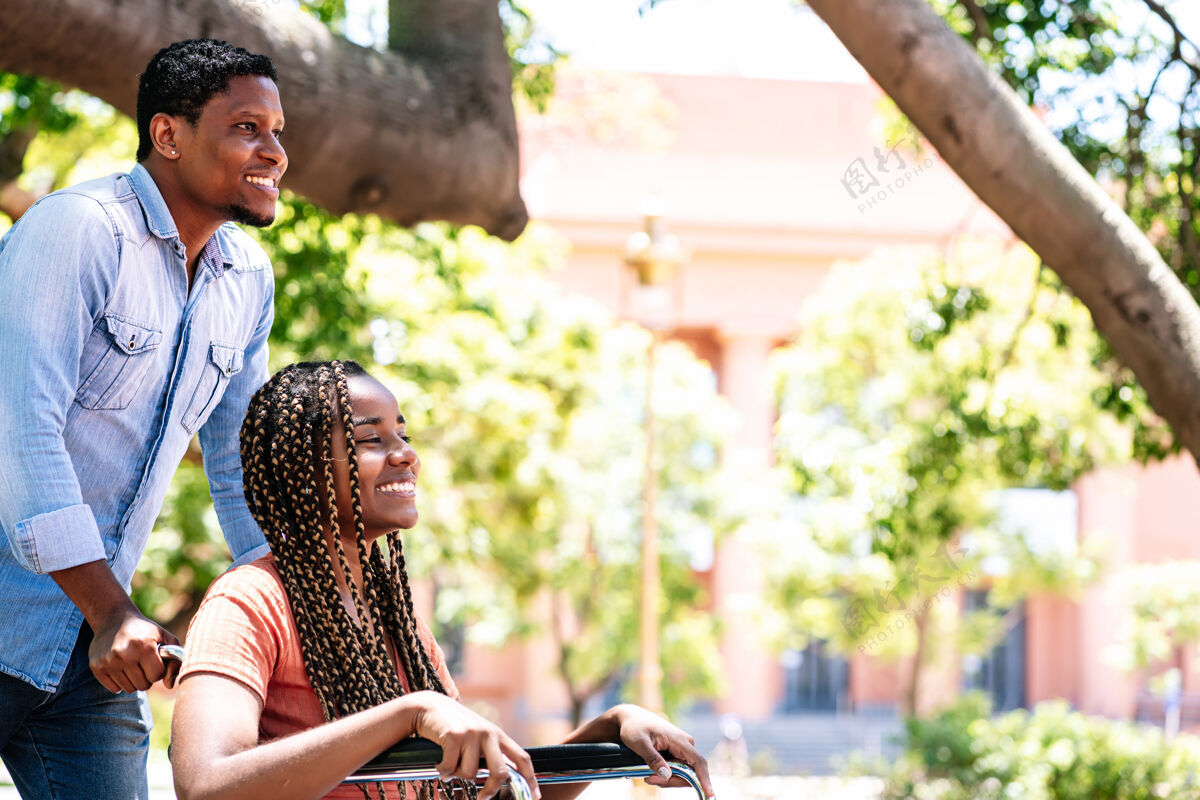 户外一个坐在轮椅上的非裔美国妇女和她的男朋友在公园散步帮助休闲消费