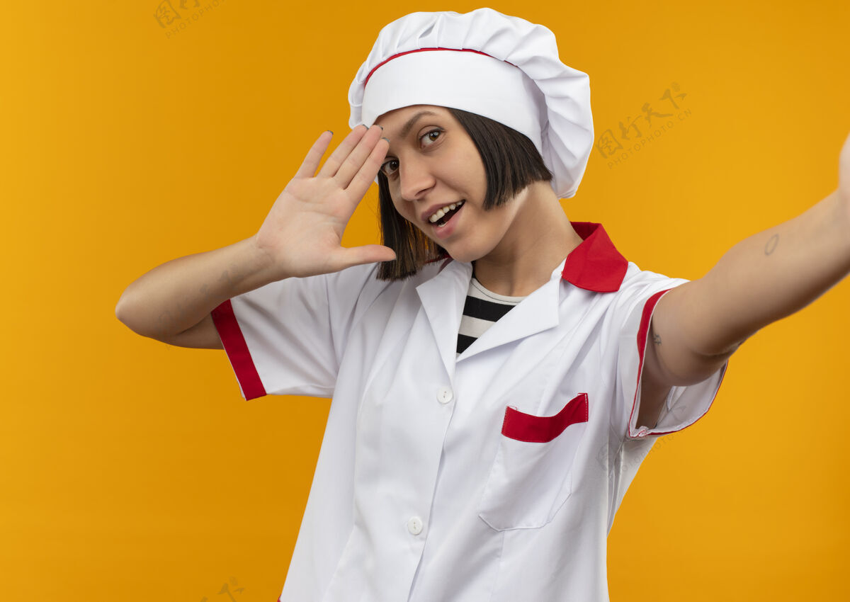 橙色身着厨师制服的快乐年轻女厨师伸出手 在橘子上打招呼你好手势女