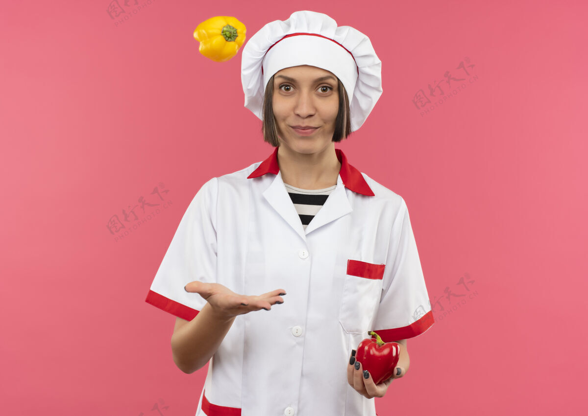 烹饪身着厨师制服的年轻女厨师手拿辣椒 把辣椒扔到粉红色背景上 并留有复印空间年轻厨师拿着