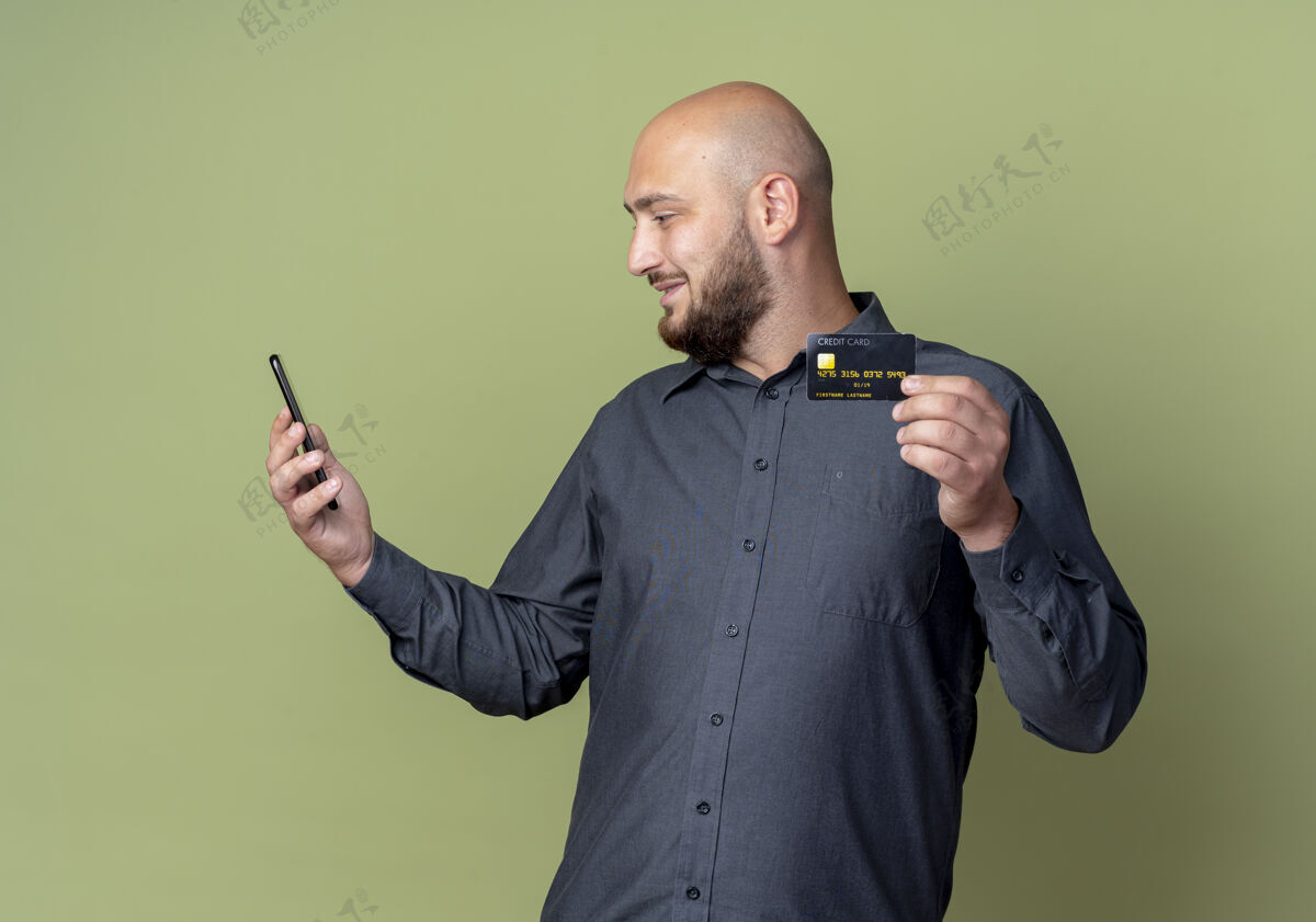 电话高兴的年轻秃头呼叫中心男子拿着手机看着信用卡在另一只手上孤立的橄榄绿背景手另一个信用