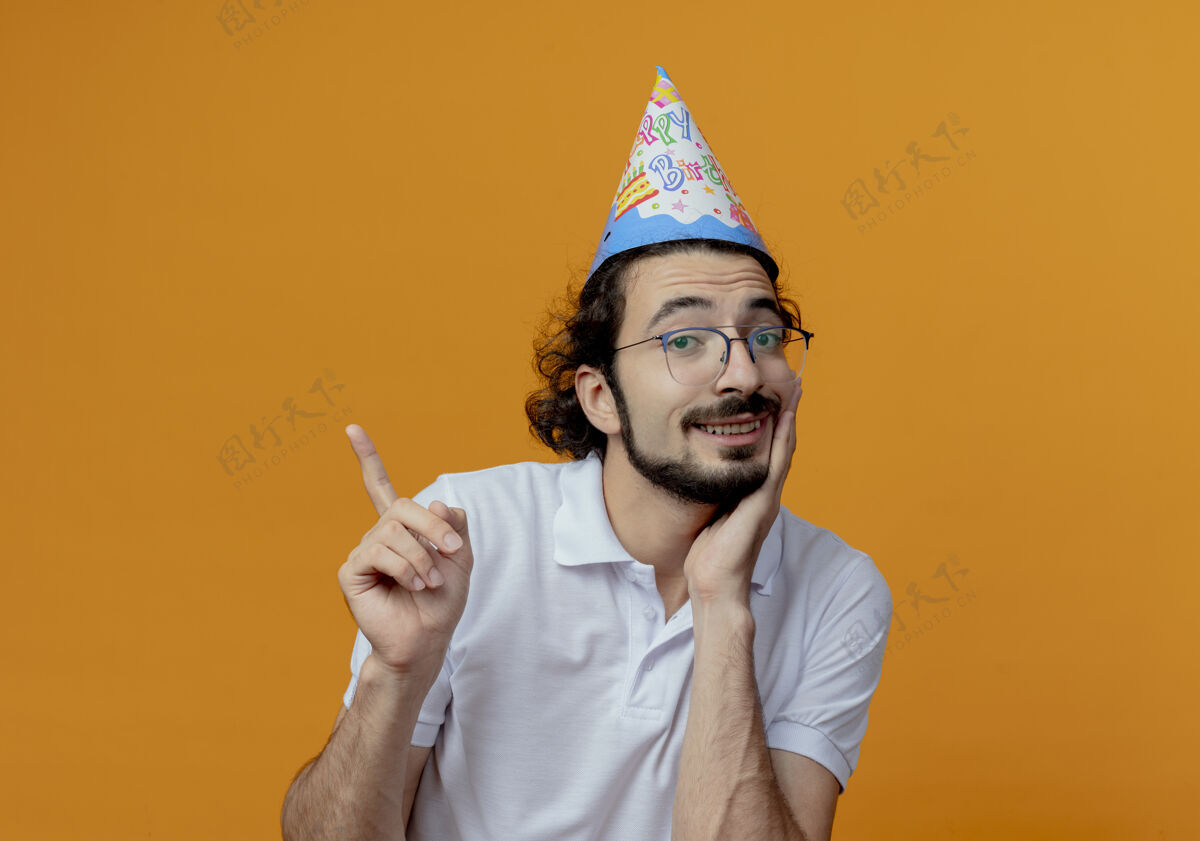 壁板笑容可掬的帅哥戴着眼镜和生日帽点在一旁 把手放在下巴下 隔离在橙色背景上 留有临摹空间眼镜尖手
