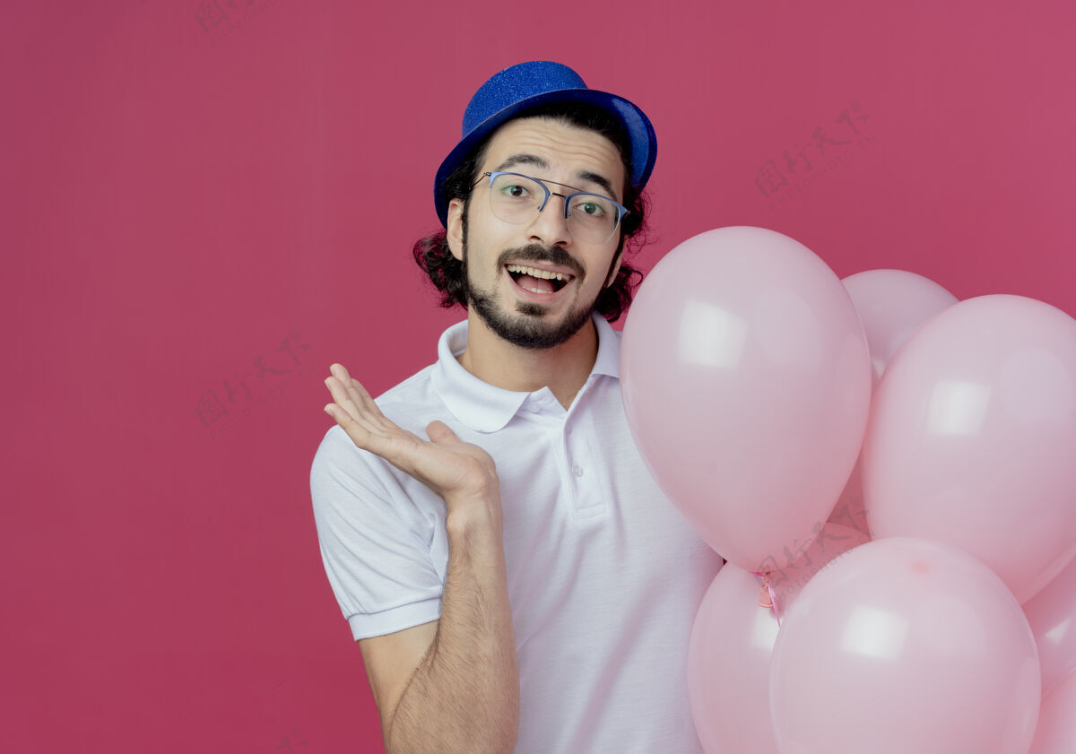 气球笑容可掬的帅哥戴着眼镜 戴着蓝色帽子 手里拿着气球 用手点着 在粉色背景上隔离开来 留有复印空间帅气眼镜手