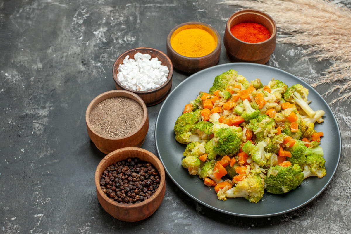 香料健康餐的侧视图 黑色盘子上有花椰菜和胡萝卜 灰色桌子上有香料胡萝卜胡椒蔬菜