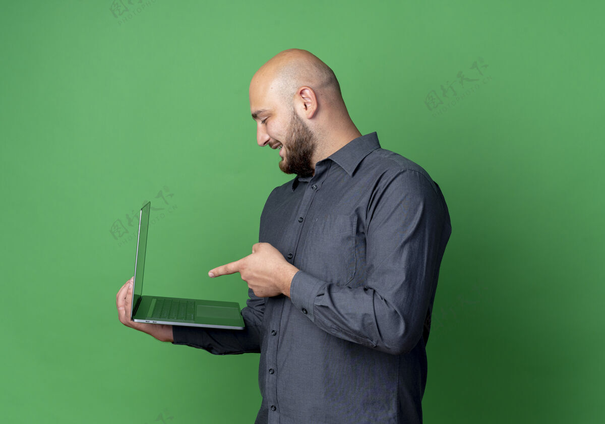 指向快乐的年轻秃头呼叫中心男子站在侧视图举行 并指着笔记本电脑孤立的绿色中心持有笔记本电脑