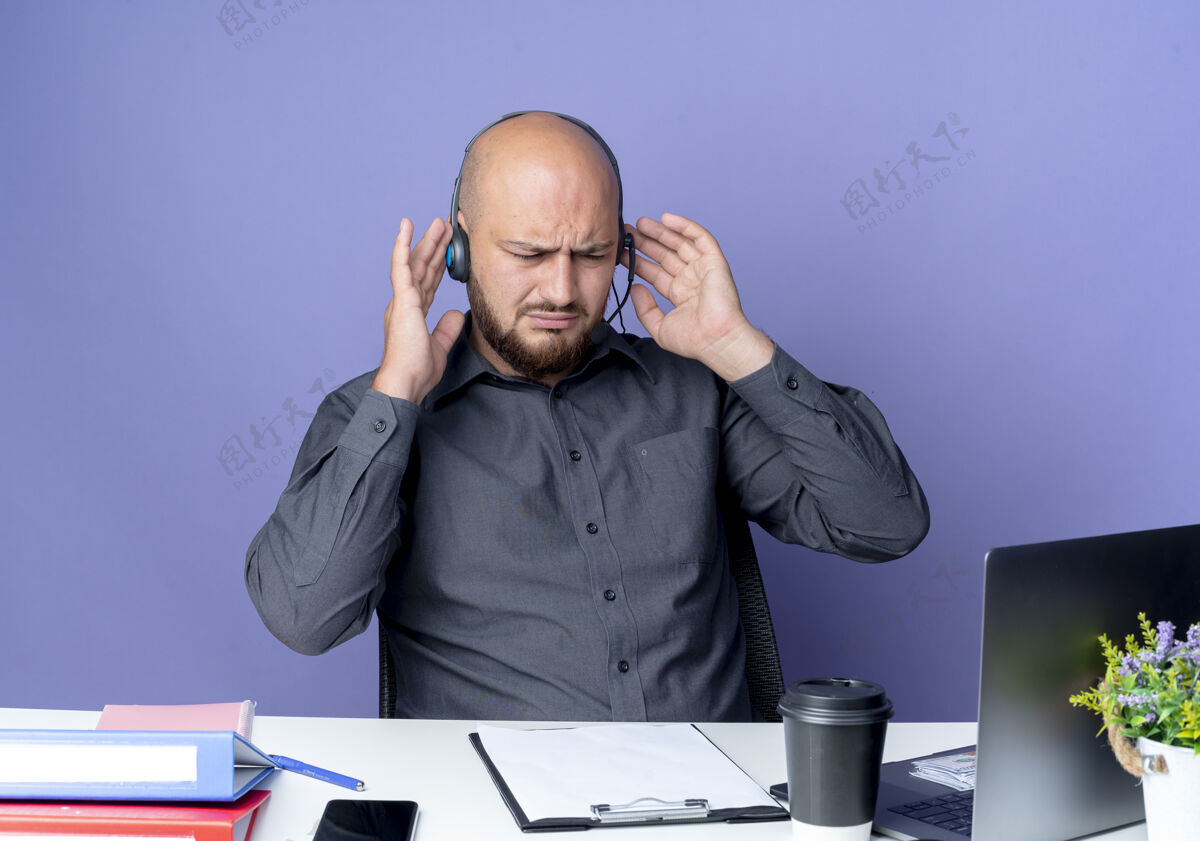 耳机戴着耳机的年轻秃头呼叫中心男子坐在办公桌旁 手放着耳机 手放在紫色背景上 低头看着工作工具工具下来坐着
