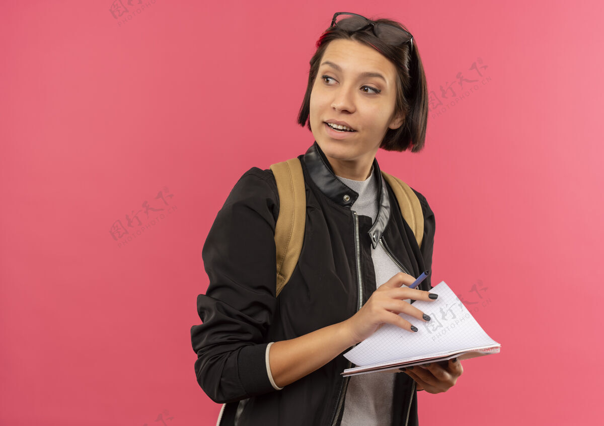粉红色印象深刻的年轻女学生戴着眼镜 头上背着书包 手里拿着笔和笔记本 一边看着粉色的隔离带壁板笔记学生