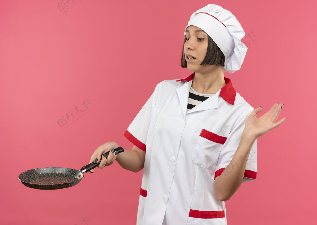 手身着厨师制服的年轻女厨师手拿煎锅看着 举起手孤立地站在粉色背景上持有厨师烹饪