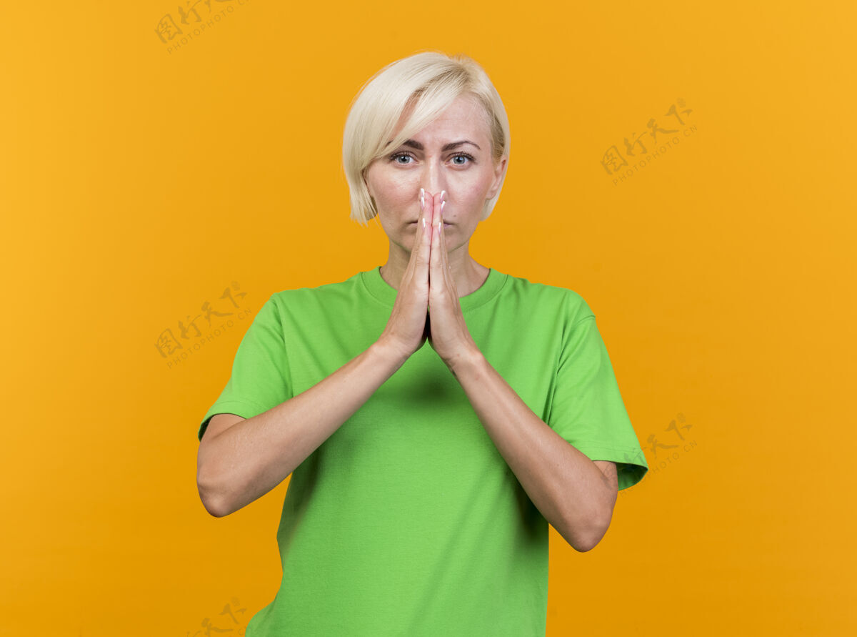 人自信的中年金发斯拉夫女人看着前方双手合十祈祷的姿势 在黄色的墙壁上隔离着嘴巴 留着复制空间手势站立女人