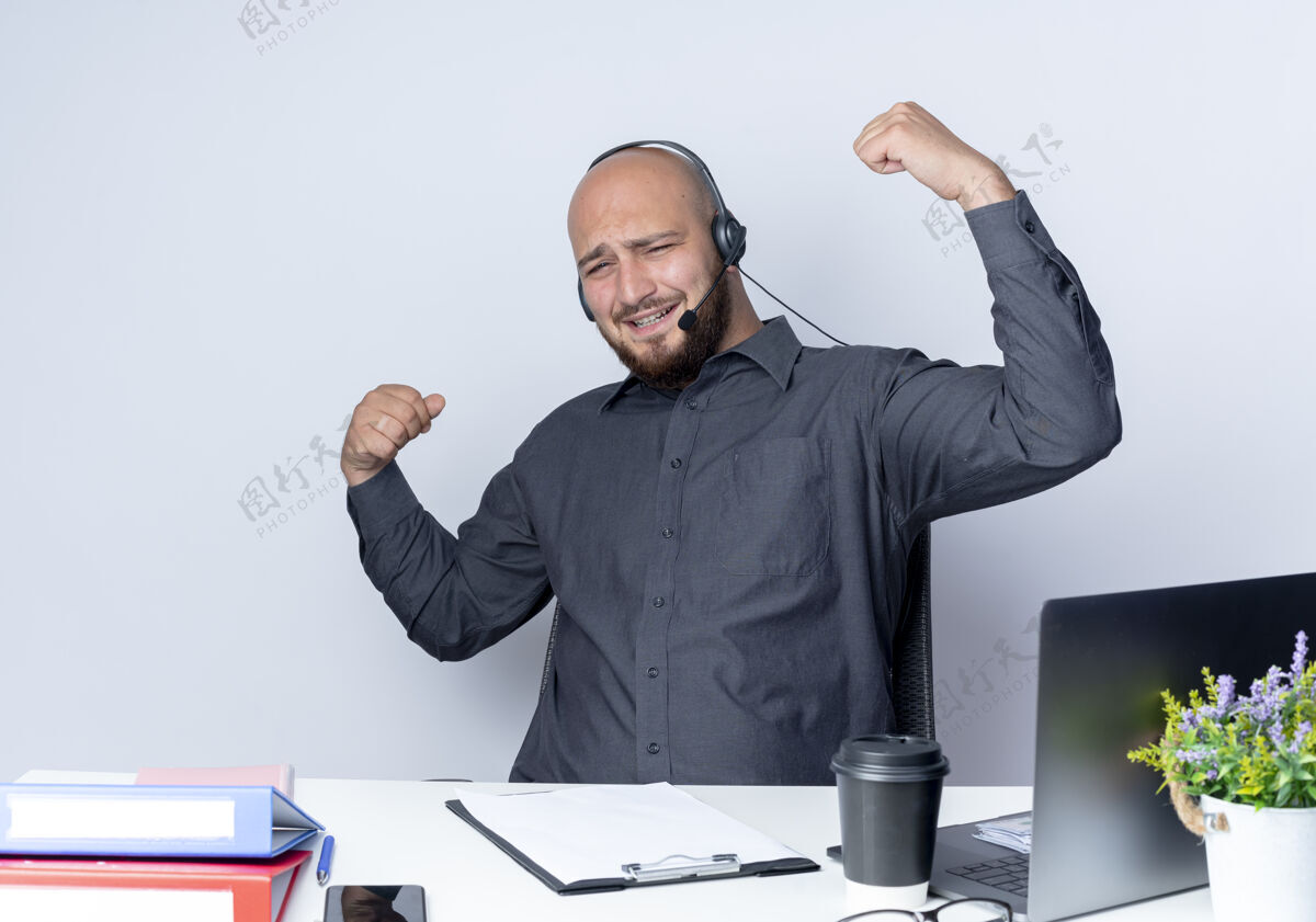 强壮快乐的年轻秃头呼叫中心男子戴着耳机坐在办公桌旁 用工作工具在白色上做着强烈的孤立的手势工具电话男人