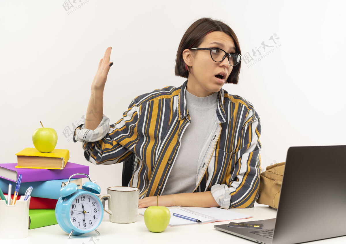 姿势不高兴的年轻女生戴着眼镜坐在桌子上 用大学的工具做手势 不看在白色背景的一边工具穿着壁板