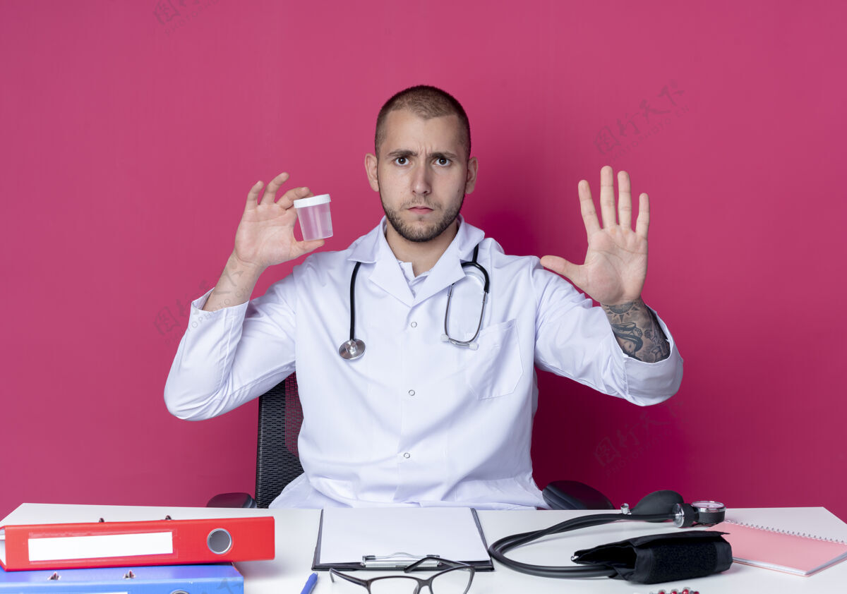 不愉快穿着医用长袍和听诊器的年轻男性医生坐在办公桌旁 手里拿着医用烧杯 五只手被隔离在粉色背景上医生工具听诊器