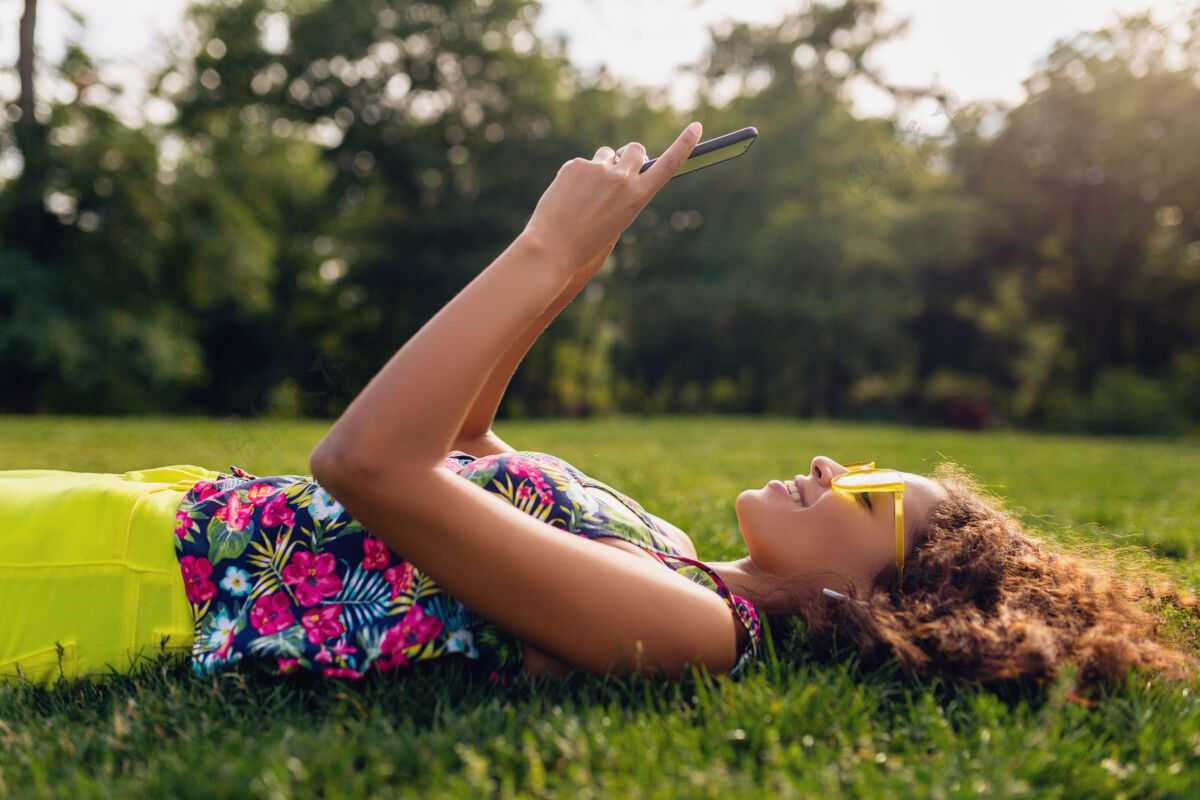 酷年轻时尚笑脸的黑人女子用智能手机听音乐无线耳机在公园里玩得开心 夏日时尚缤纷风格 躺在草地上 戴着黄色太阳镜享受混血儿黑色