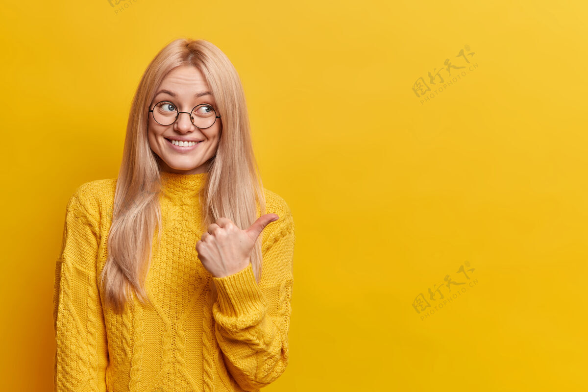 微笑可爱的金发女郎微笑着 指着黄色墙上的复制空间年轻手指解决方案
