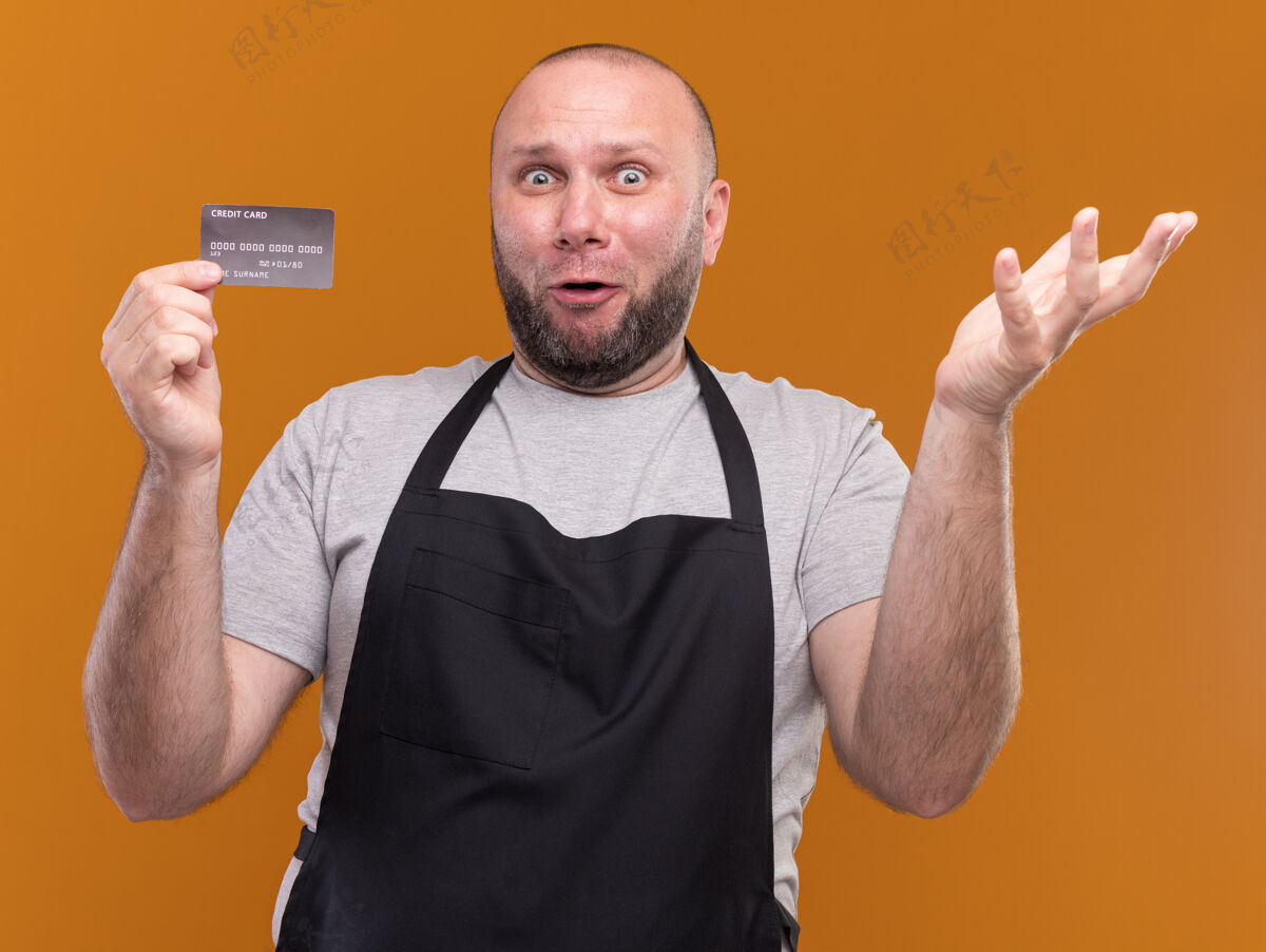 持有惊讶的斯拉夫中年男理发师穿着制服拿着信用卡摊开的手孤立在橙色的墙上蔓延中年斯拉夫人