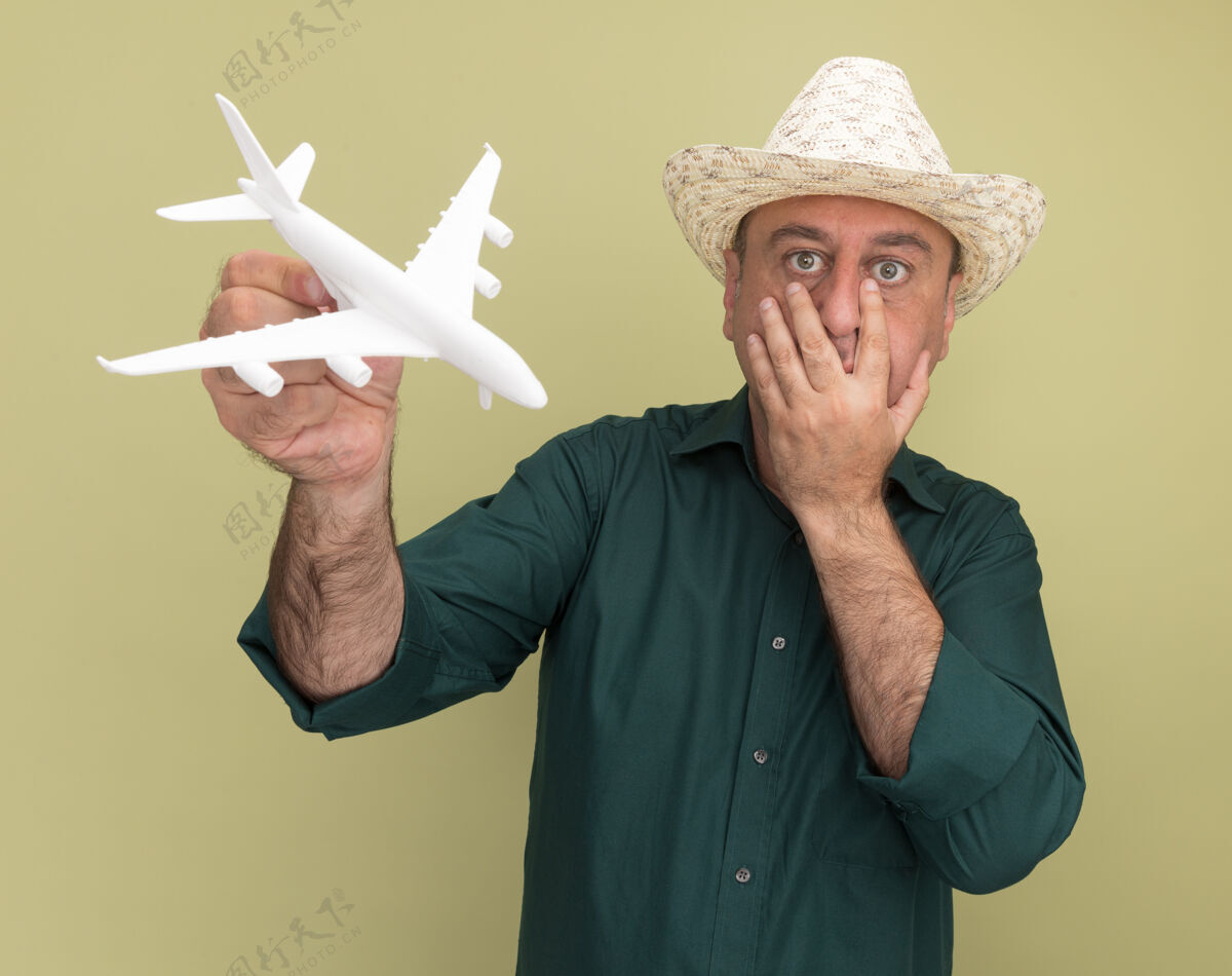 帽子惊恐的中年男子穿着绿色t恤 戴着帽子 手里拿着玩具飞机 手放在嘴上 隔离在橄榄绿的墙上穿橄榄T恤