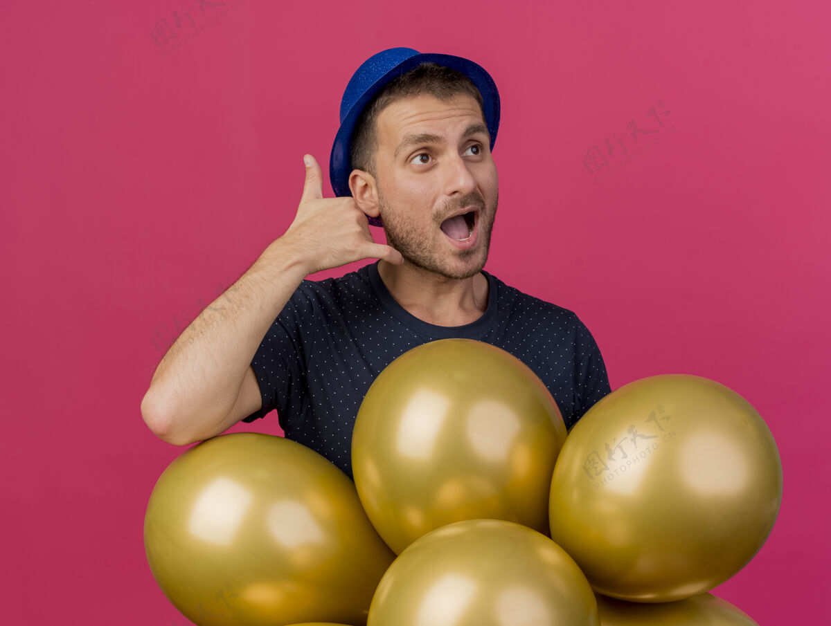 气球快乐英俊的高加索男子戴着蓝色派对帽手势叫我的标志 拿着氦气球看着粉红色背景上的一面 并留有复制空间复制蓝色手势