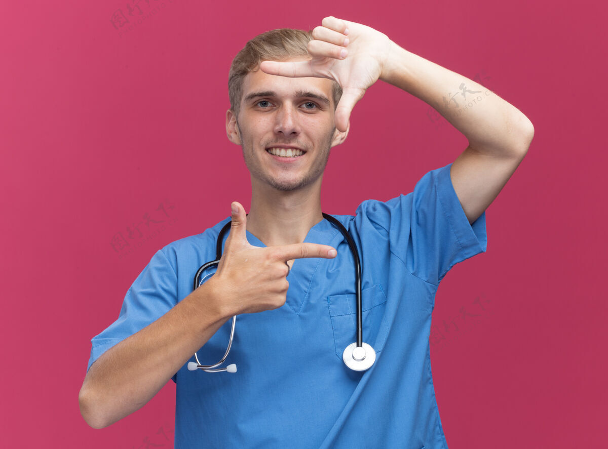 佩戴微笑的年轻男医生穿着医生制服 带听诊器 在粉红色的墙上显示照片姿势男性年轻手势
