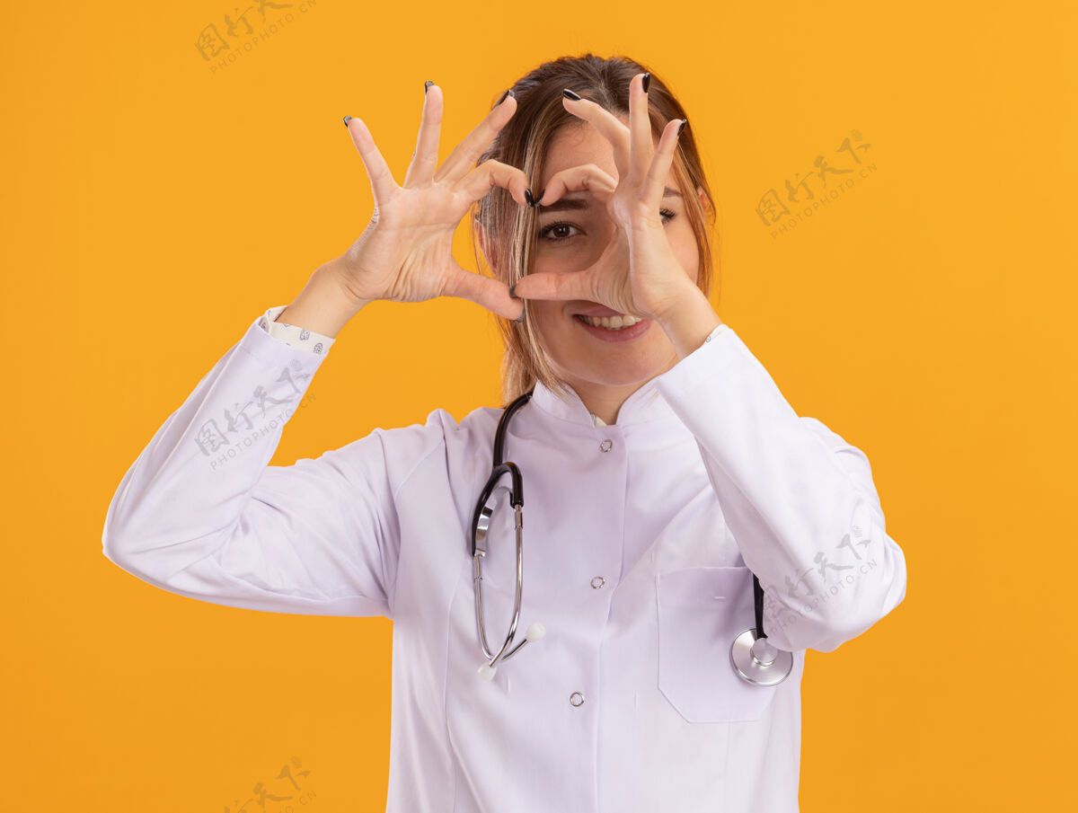 表演面带微笑的年轻女医生 穿着医用长袍 听诊器显示出孤立在黄色墙上的心脏姿势长袍医学年轻人