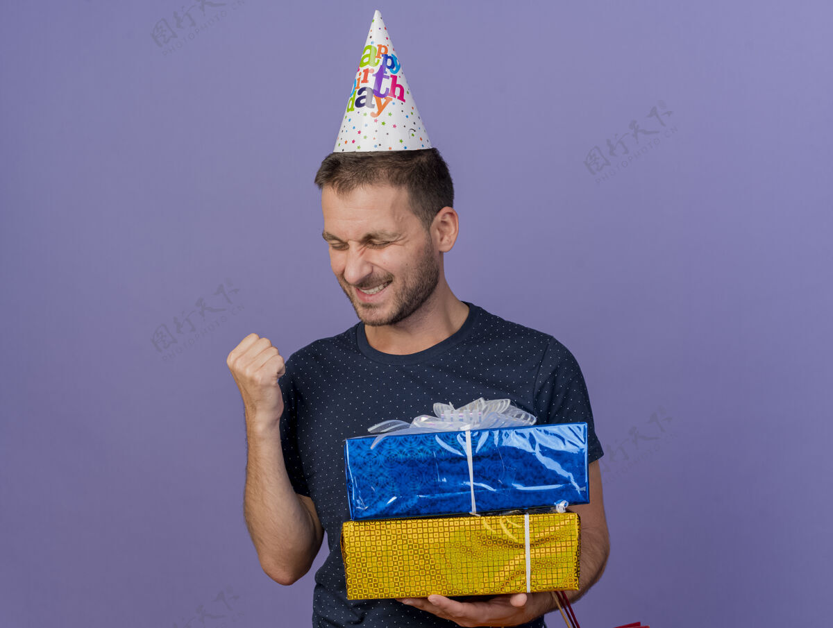复制戴着生日帽的快乐帅哥高加索人握紧拳头 拿着紫色背景上的礼品盒 还有复制空间帽子持有男人