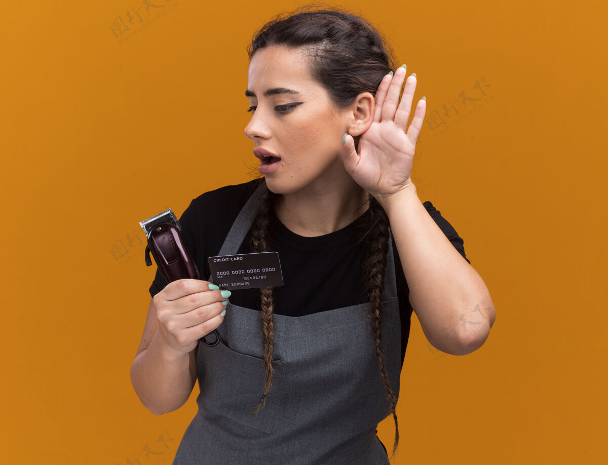 年轻可疑的年轻女理发师穿着制服 手里拿着信用卡和理发器 在橘黄色的墙上显示着被隔离的姿势显示手势制服