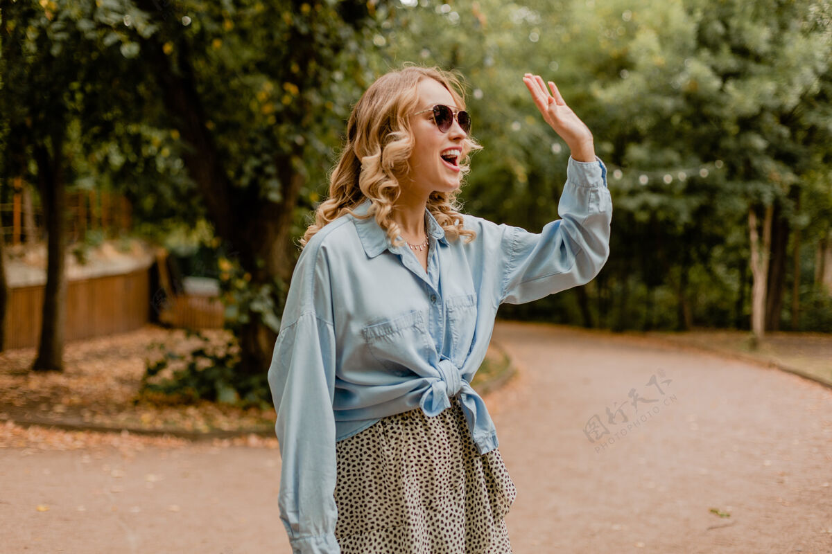 衣服迷人的金发微笑的女人挥手问候在公园散步在夏天的服装太阳镜欢呼时尚