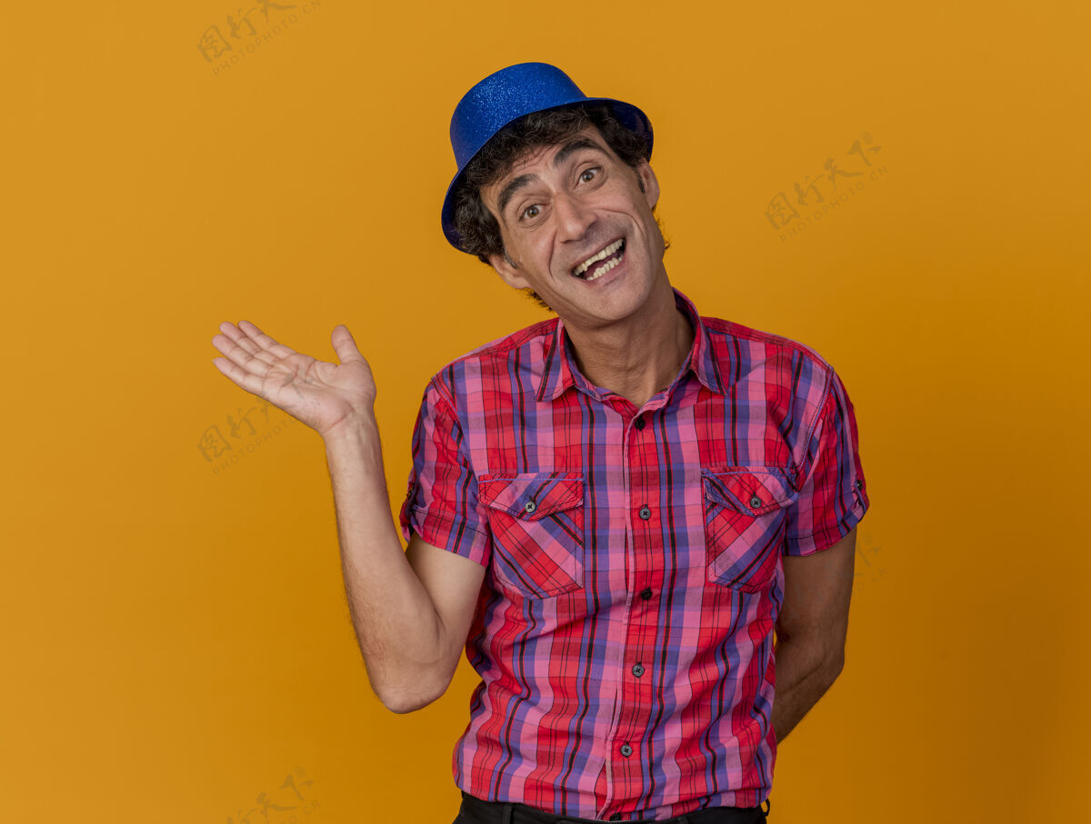 帽子快乐的中年白种人党男子戴着党的帽子看着相机显示空手保持另一个背后的橙色背景与复制空间隔离欢乐中年空