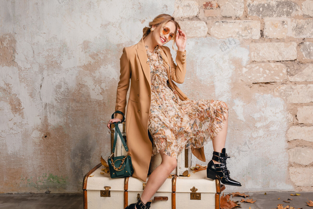 时尚在街上 穿着米色外套的金发美女坐在手提箱上对着墙衣服模特包