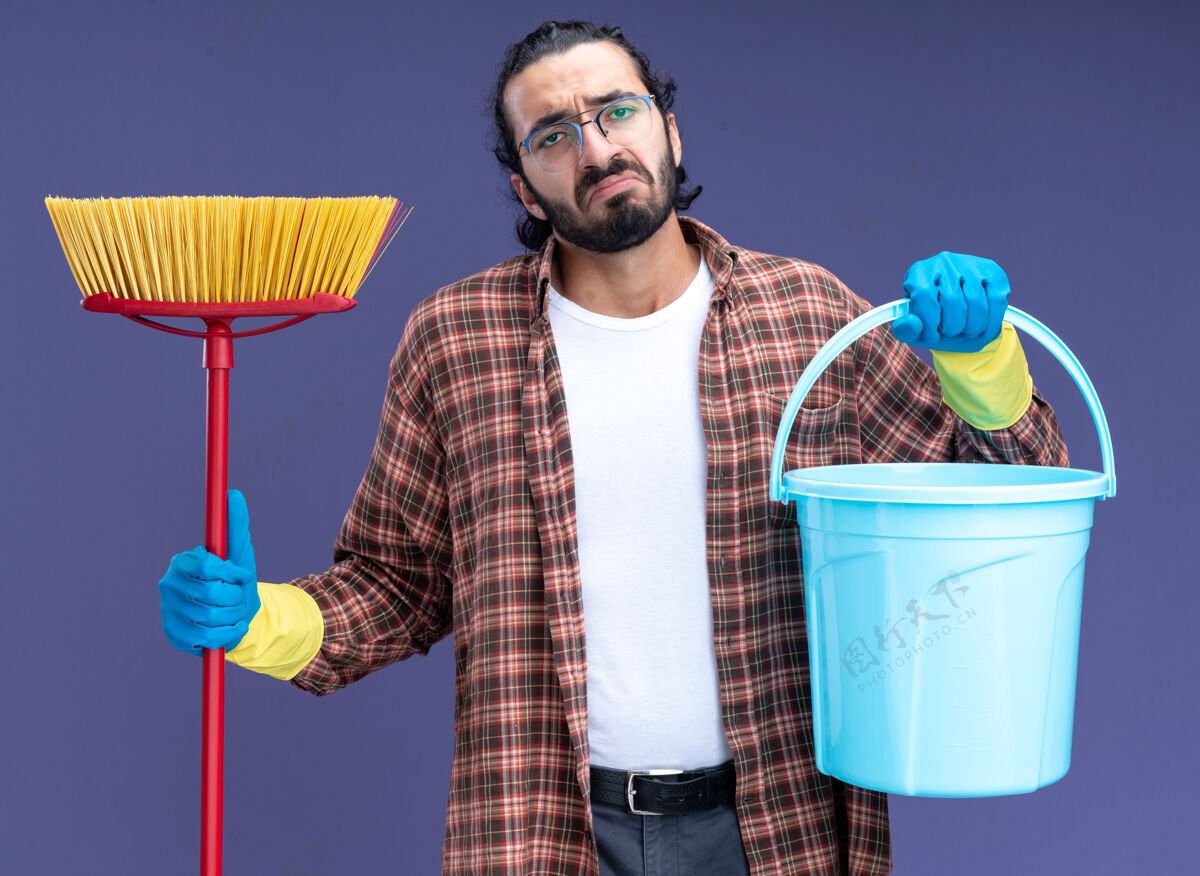 手套悲伤的年轻帅气的清洁工 穿着t恤 戴着手套 拿着水桶 把拖把隔离在蓝色的墙上清洁穿家伙