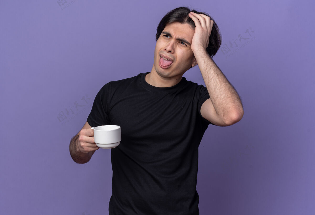 舌头穿着黑色t恤的年轻帅哥 拿着一杯咖啡 露出舌头 把手放在头上 孤立在紫色的墙上年轻人T恤杯子