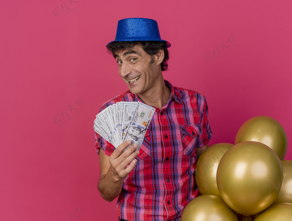 举行微笑的中年白种人党男子戴着党的帽子拿着气球和钱看着摄像机隔离在深红色的背景与复制空间男人金钱复制