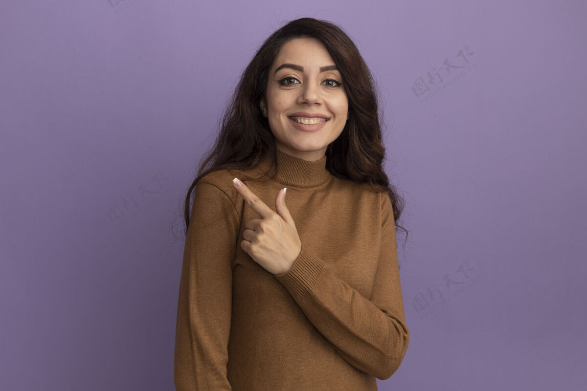 毛衣微笑的年轻漂亮女孩穿着棕色高领毛衣点在一边孤立的紫色墙壁与复制空间年轻微笑壁板
