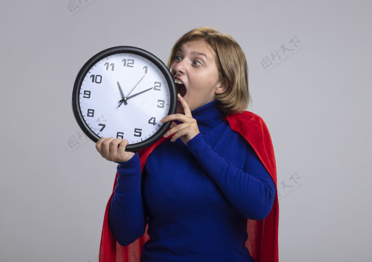试试年轻的金发超级英雄女孩在红色斗篷举行 并看着时钟试图咬它的白色背景与复制空间隔离超级英雄抱着太空