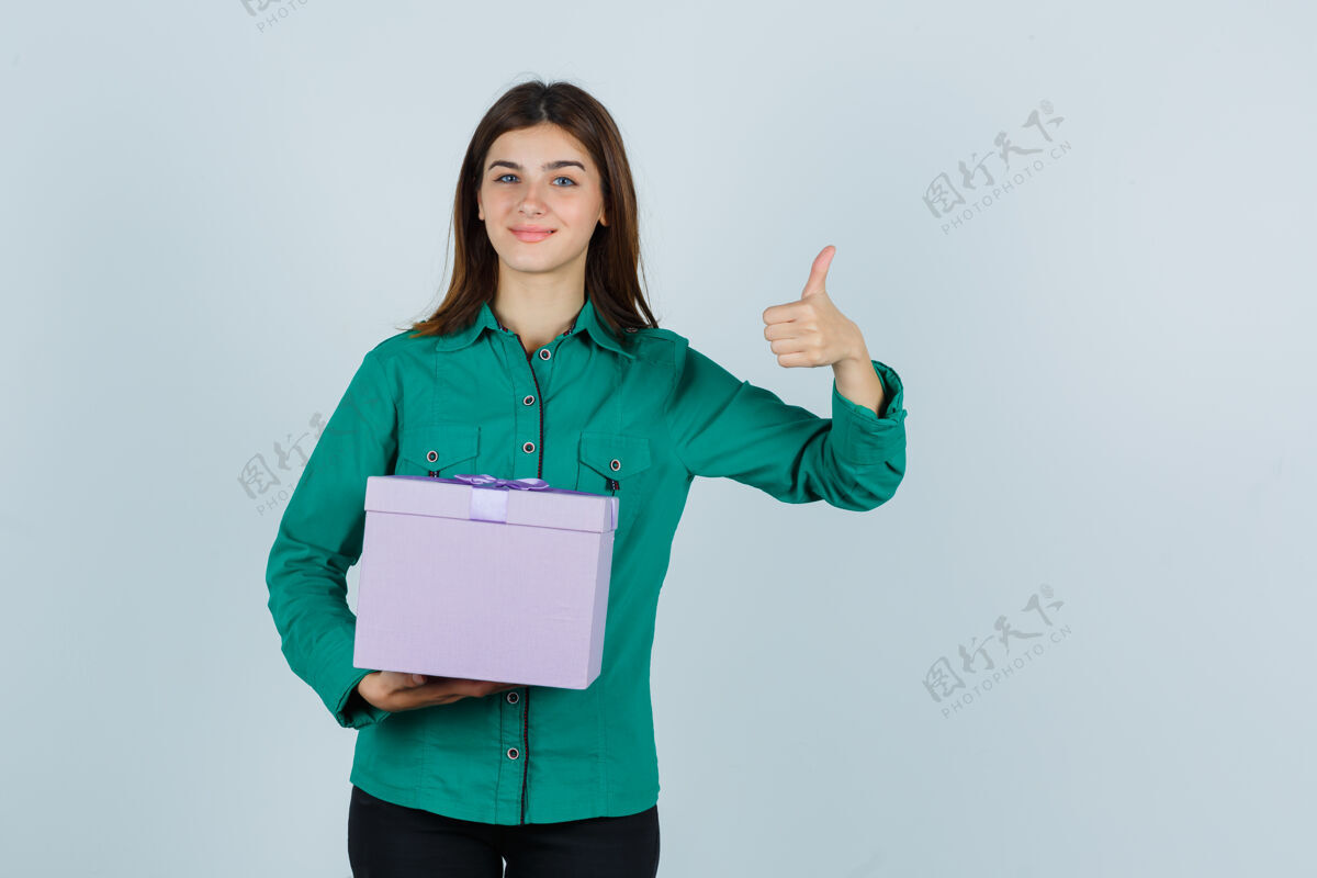 积极拿着礼盒的年轻女孩 穿着绿色上衣 黑色裤子 竖起大拇指 看起来很快乐 正前方的景色拇指青少年女性