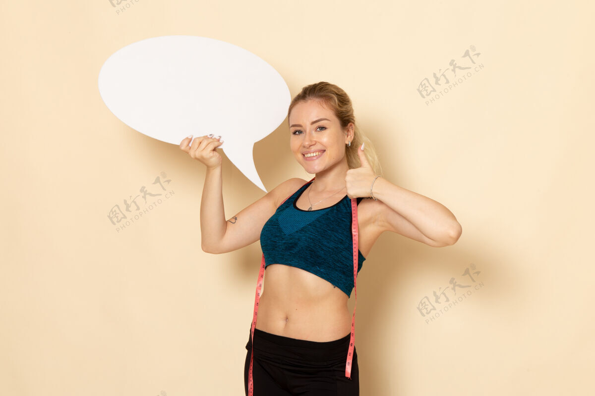 运动正面图身穿运动装的年轻女性手持白色标志 在白色墙上测量身体运动美容健康锻炼适合健身测量装备