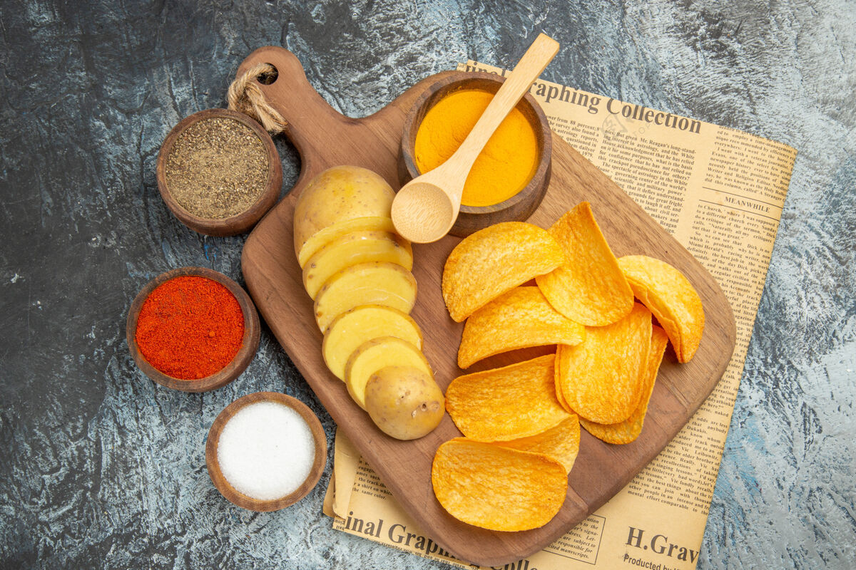 餐上图：美味的自制薯片放在木制砧板上切土豆片 不同的香料放在灰色桌子上的报纸上食物饮食土豆