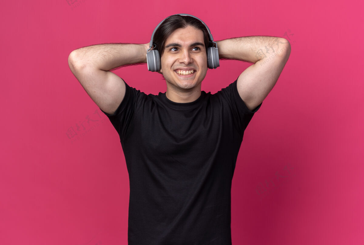 耳机面带微笑的年轻帅哥穿着黑色t恤 戴着耳机 把双手放在头后 隔离在粉红色的墙上年轻人头微笑
