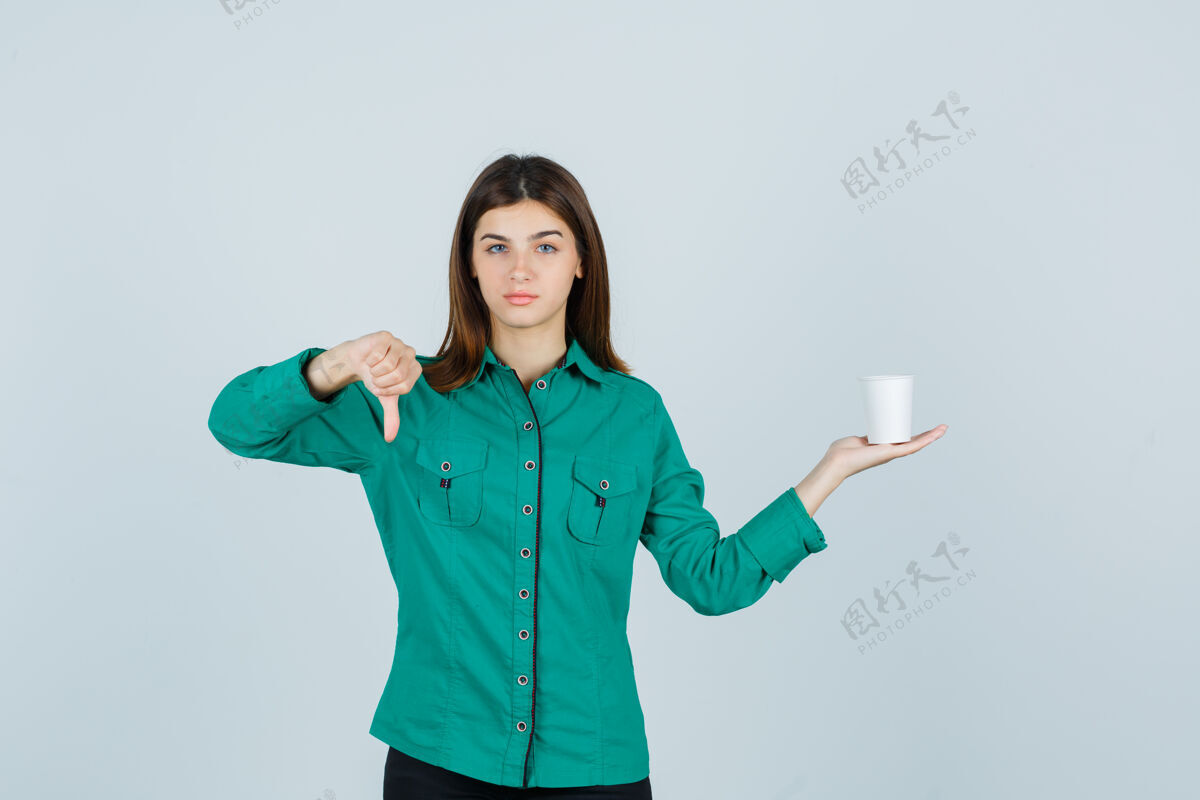 成人年轻的女士拿着一杯塑料咖啡 大拇指朝下 穿着衬衫 看起来很不满意 正面图眼睛年轻焦点