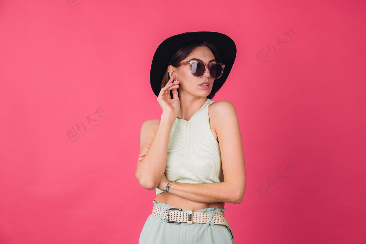 模特戴着帽子和太阳镜的漂亮时髦女人在粉红色的墙上摆姿势嘴唇肖像衣服