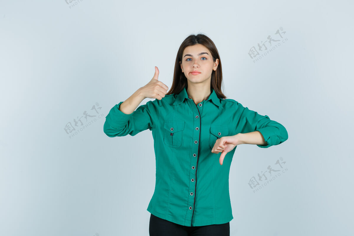 前面年轻的女性展示相反的拇指 弯曲的嘴唇在绿色衬衫 看起来优柔寡断前视图人女性优柔寡断