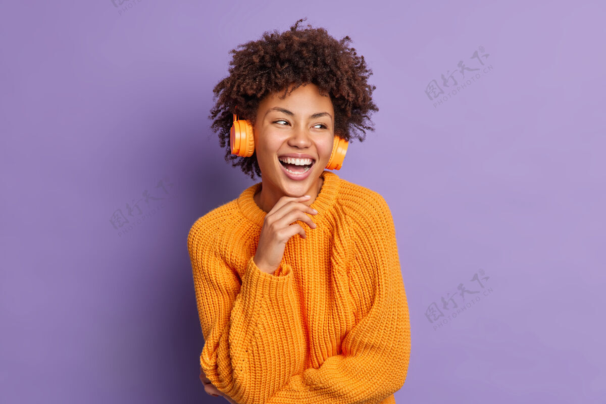 表情正黑皮肤的年轻女子戴着耳机听音乐 抱着下巴 微笑着高兴的专注在一旁 喜出望外地穿着暖和的针织橘色套头衫乐观种族欣喜若狂